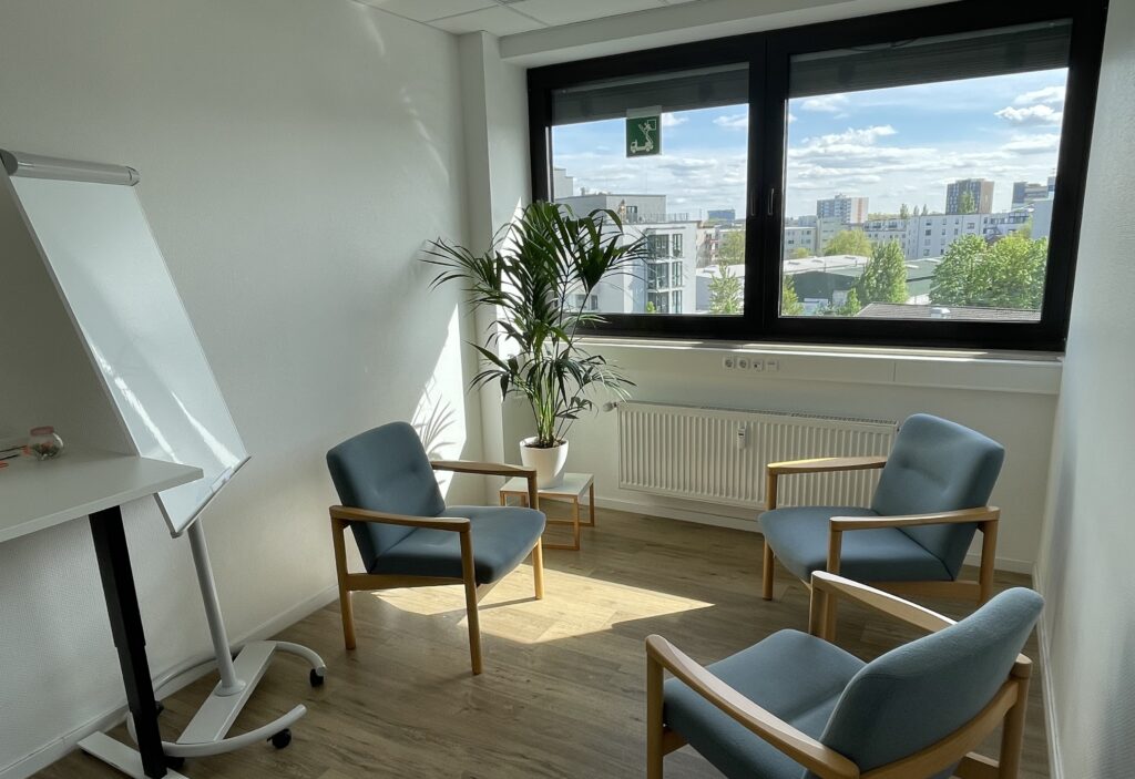 Zu sehen ist der Raum, in dem das Coaching stattfindet: 3 Stühle aus Holz mit blauer Polsterung, die im Dreieck einander zugewandt stehen. Dahinter sieht man das Fenster und den Stadtblick aus der 4. Etage. Links neben den Stühlen steht eine Flipchart.